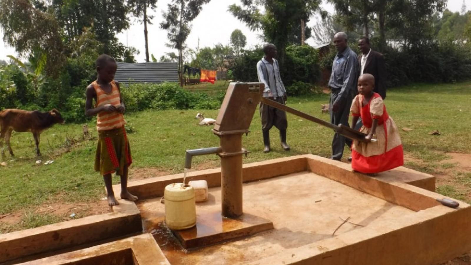 A group of black people, three adults and two children, standing around a water pump. One of the kids is pumping water into a plastic jug.