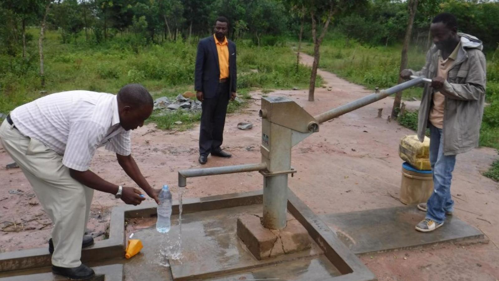 Three black men around a bump. One is pumping water, another is putting a plastic water bottle under the spout.