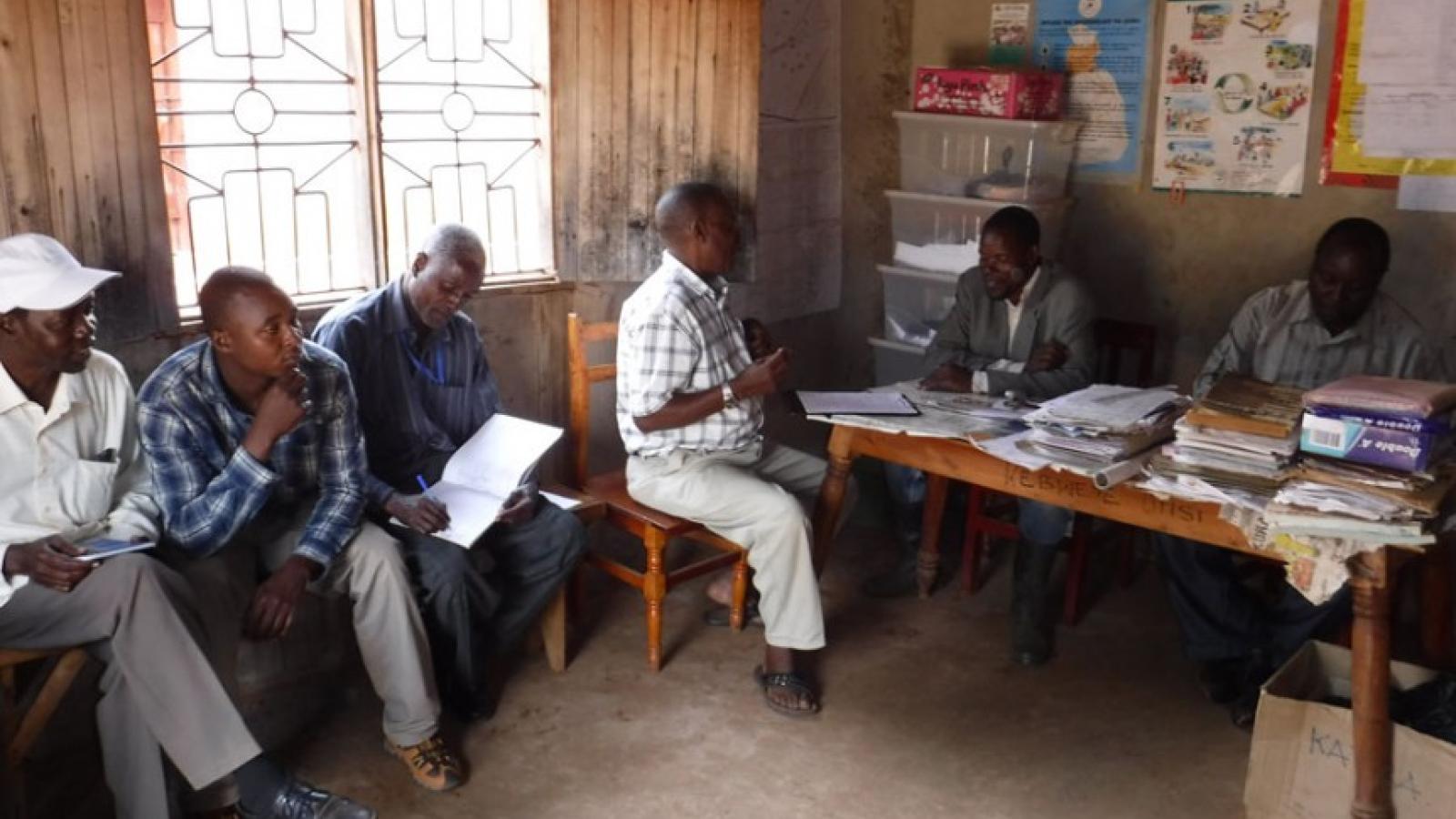 Inside a dirt-walled building with decorated walls and windows, six black men are sitting in a meeting. Two are behind a table covered in papers and books. The others sit in chairs along the wall.