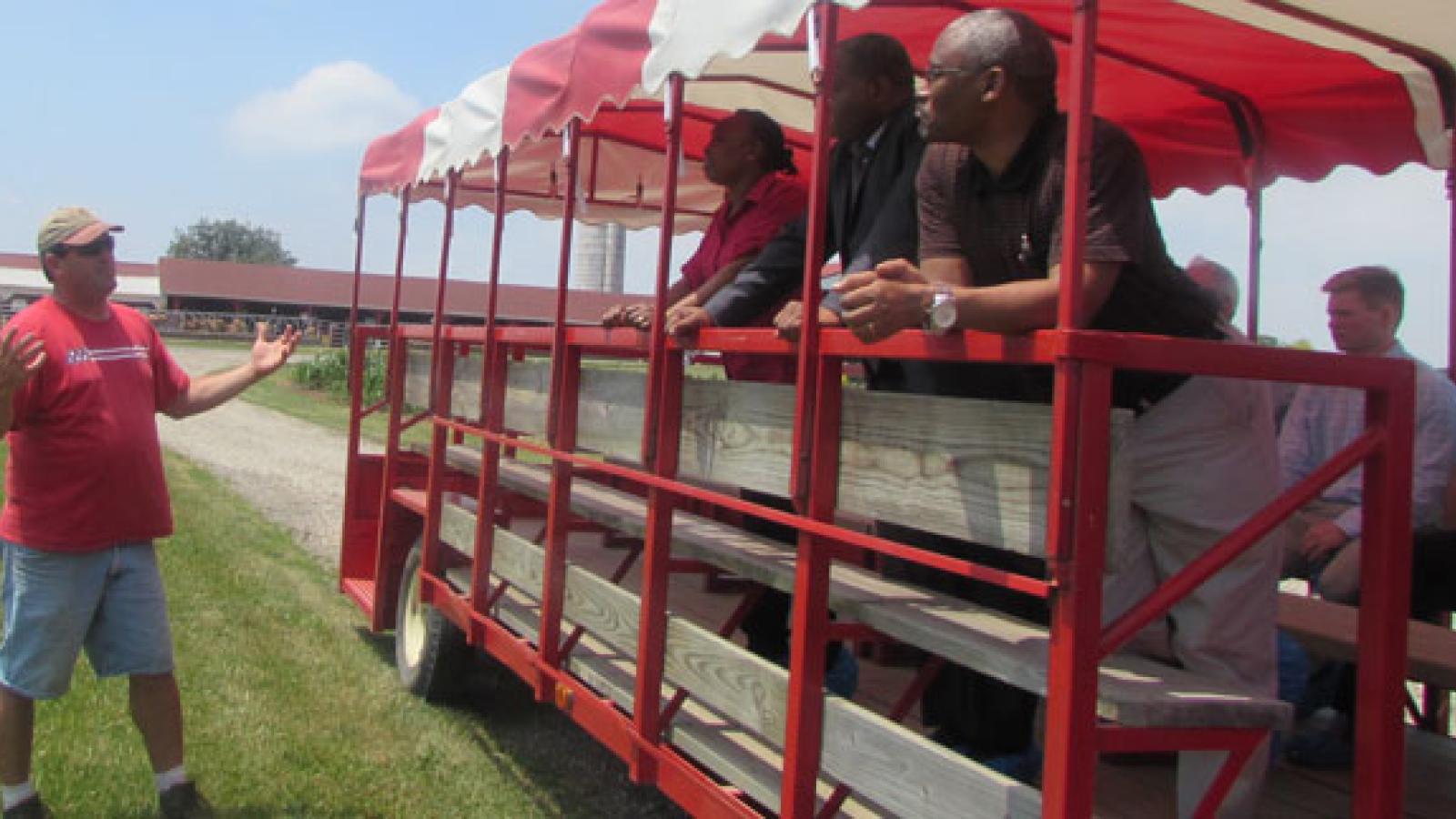 A group of people standing in a trailer that had benches and an red/white stripped canopy. Outside the trailer is a white man in a red tee shirt. He is mid-gesture and seems to be in conversation with a few of the people on standing in the trailer