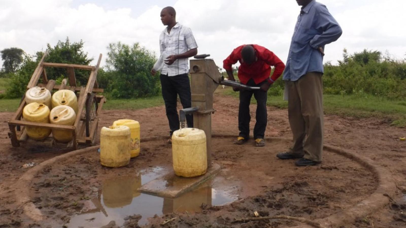 Three black men standing around a water pump. One of the men is pumping water into a plastic jug. Nearby are two other jugs on the ground and a cart with four stacked jugs