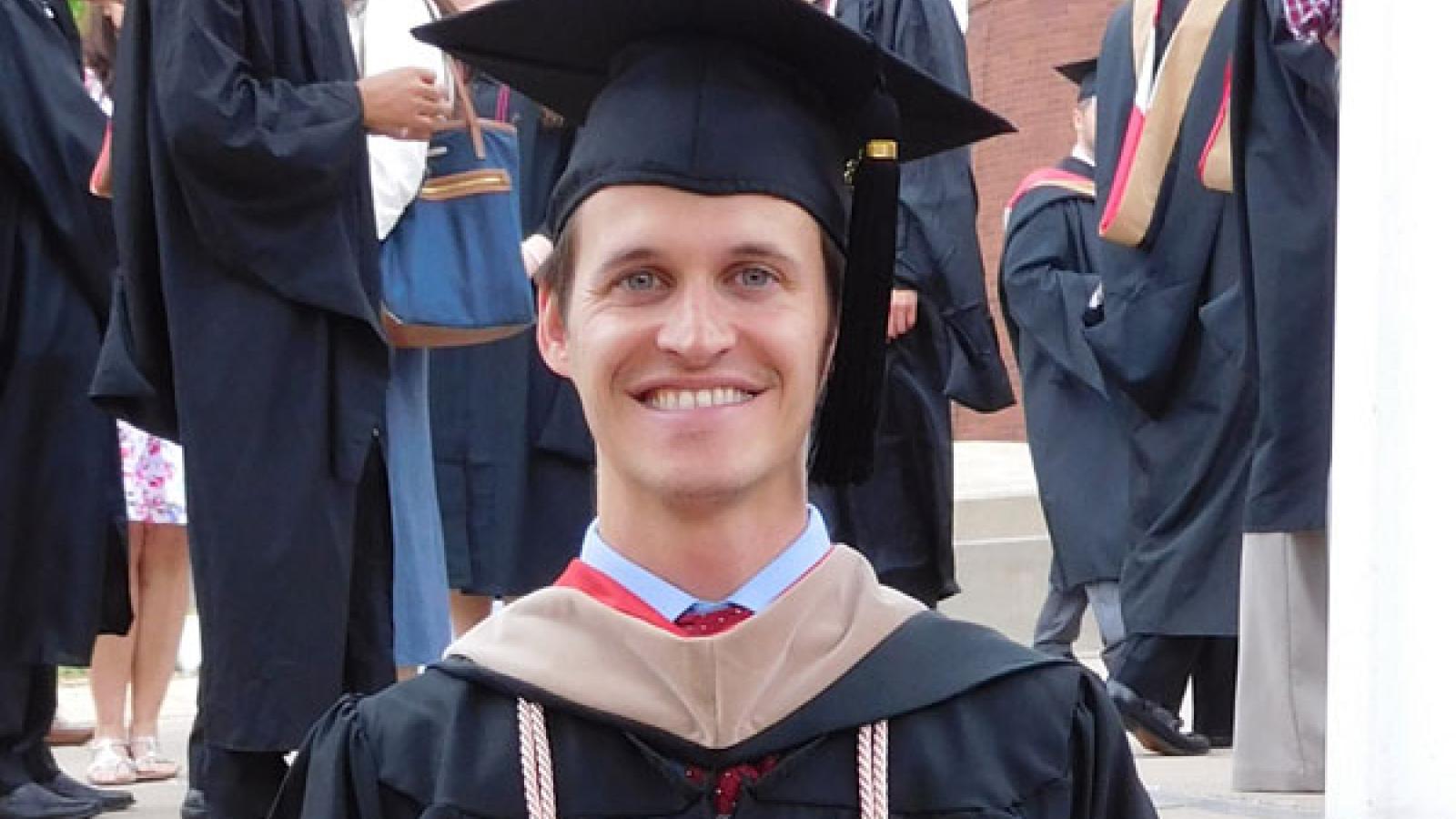 A white man in a graduation cap and gown smiling at the camera