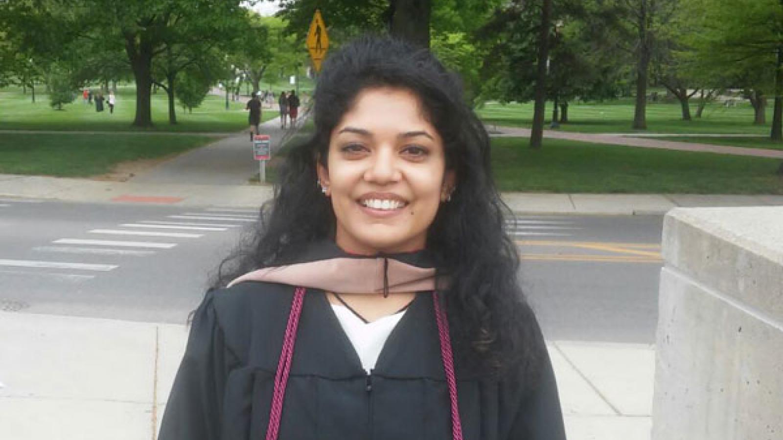 A woman with black, curly hair and light brown skin. She is wearing a graduation gown and smiling.