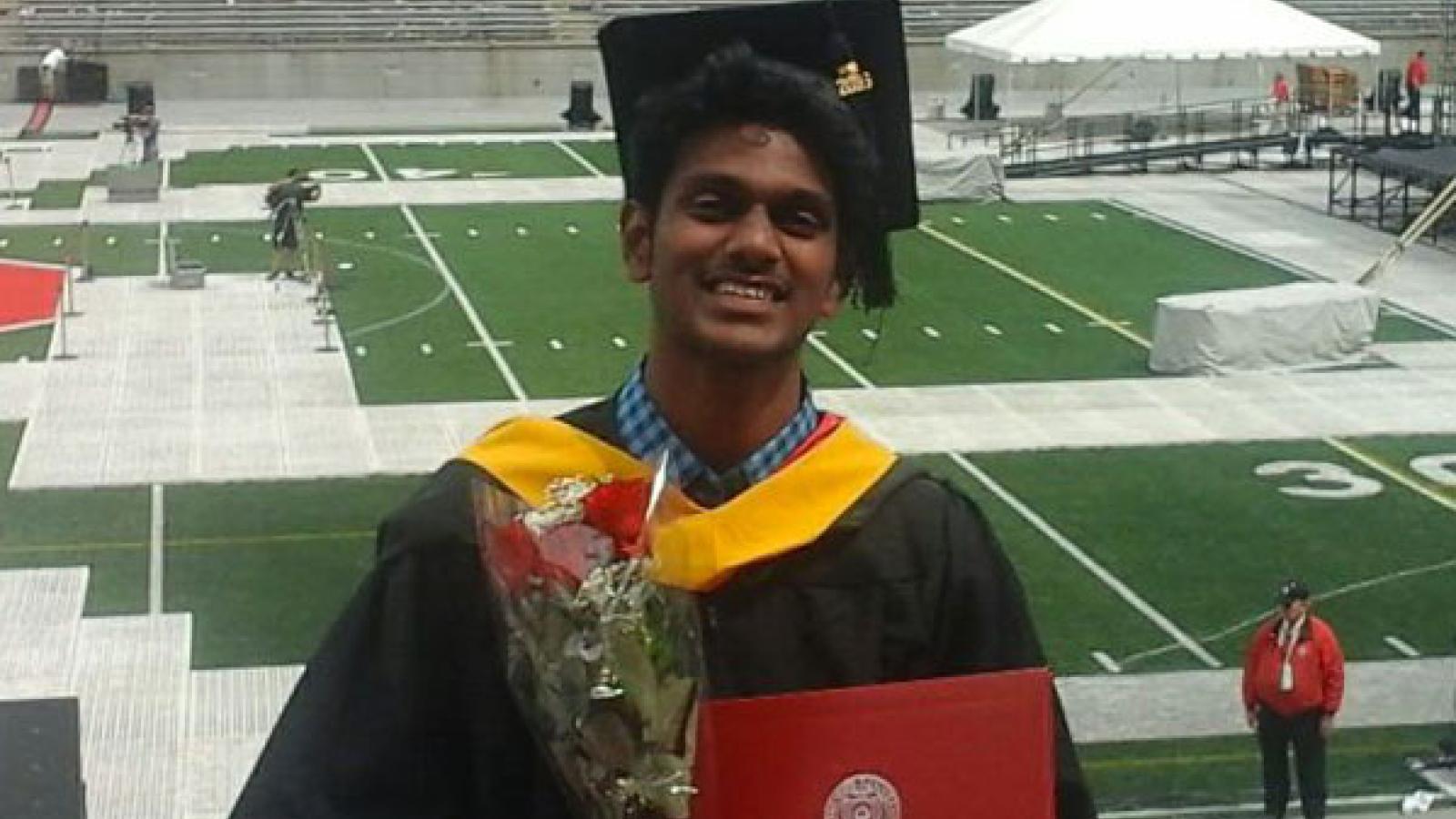 An Indian man with dark skin and black hair smiling at the camera. He is wearing a graduation cap and gown, and holding a bouquet of roses and a diploma