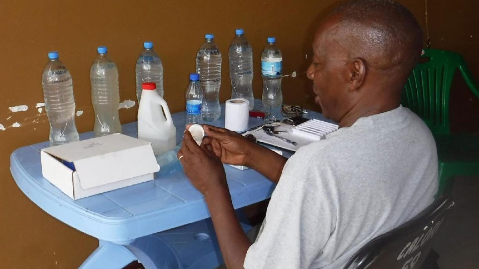 A bald, black man in a white tee shirt sitting at a table. On the table are water bottles, a jug, a box, a roll of toilet paper, keys, and a clip board with some paper 