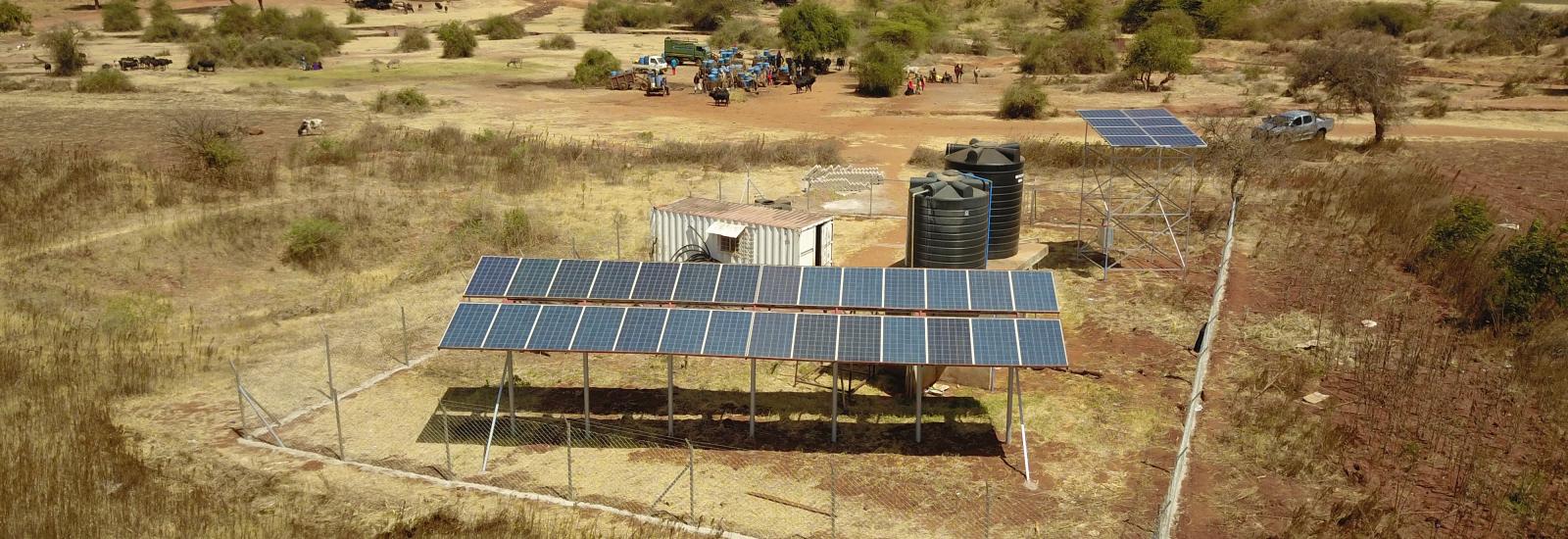 2 sets of solar panels perched up on metal frames fenced in with wire fence with 2 large black water cylindrical water reservoirs and a white trailer surrounded by dirt, dry grass and trees with people and animals in a distance. 